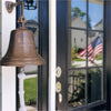 8 inch diameter distressed, antiqued finish solid brass wall bell with two lines of custom engraving hanging near front door of porch with American flag reflected in glass