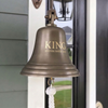 Antiqued brass 10 inch diameter bell mounted on a wall near a front door with the words "KING" and "Established 2024" engraved on the front