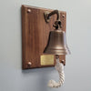 A bronze bell mounted on a rectangular wooden plaque with brass screws and a brass nameplate, hanging against a light gray wall. A white rope knotted below the bell is visible.