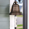 6 inch diameter antiqued brass wall bell with knotted pull rope mounted on a wall near a front door withe 3 lines of engraving