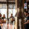 7 inch diameter antique finish brass wall bell mounted on a wood post on a restaurant bar