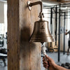 7 inch antiqued finish wall bell mounted on a rough vertical beam in a gym with words PR BELL engraved on the face of the bell and a hand pulling the knotted pull rope
