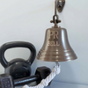 7 inch antiqued finish brass wall bell on a light blue grey background with cast iron weights in the background