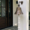 A bronze-colored metal bell mounted on a white exterior wall beside a dark wood front door. The bell is engraved with a large letter C and the phrase 'Welcome to Our Home The Crooks', and hangs with a white knotted rope.