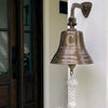 A bronze outdoor bell mounted on a white wall with a dark wooden door to the left. The bell is engraved with a large letter C and the words 'Welcome to Our Home The Cooks', and hangs from a white rope.