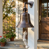 A bronze-finished brass bell with engraved text reading 'Lake House Established 1917 Austin, Texas' hanging from a  bracket on a white porch post, with a knotted white rope below and a wooden front door in the background.