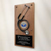 A wooden wall plaque featuring a black stethoscope mounted on a brown wood panel, with a round medical badge and a black engraved plaque at the bottom containing a quote.