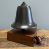 A dark brass bell mounted on a rectangular wooden base, with a small wooden gavel resting against the side of the base, set on a rustic wooden surface against a plain gray wall.