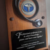 A wooden medical graduation plaque featuring a black stethoscope and a circular silver-and-blue medallion with a caduceus symbol. A black inscription plate below contains a physician quote and graduation recognition text for Vanderbilt University School of Medicine.