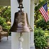 A bronze-colored metal bell hanging from a white porch post, featuring the engraved text 'WEISKE HOME & WHERE WE PUT OUR HEADS' with a mountain silhouette and oak leaves. A white rope hangs from the bottom, and an American flag is visible in the green garden background.