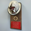 A vintage white firefighter helmet mounted on a wooden plaque with a red prayer plaque and a gold emblem, displayed against a light gray wall.