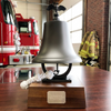 A large brass bell on a wooden base with a rope and eagle topper, displayed in a fire station with a red fire truck and firefighter jackets in the background.