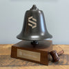 A dark brass
 bell mounted on a wooden base with a small wooden gavel, photographed indoors against a light gray wall and rustic wood tabletop. The bell and base are engraved with the Symbridge name and logo.