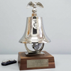 A polished nickel plate brass bell mounted on a dark wood base with a brass plaque reading 'Brooklyn Park Fire Department Est. 1965.