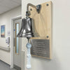 A nickel finish brass bell with a white rope pull hangs on a light wood plaque in a clinical hallway. The plaque has engraved text and is mounted on an off-white wall.