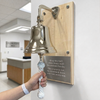 A polished brass hospital-style celebration bell with a white rope handle mounted on a wooden plaque on a white wall. A person's hand is holding the rope handle, with a hospital lobby visible in the background.