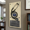 A wooden medical plaque mounted on a wall, featuring a black stethoscope and a round blue-and-white medical badge in the center, with a dark quote plaque at the bottom. The scene appears to be an office interior with framed certificates, a plant, and a bookshelf in the background.