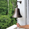 10 inch antiqued brass wall bell hanging outside of house with hand on pull rope to show size