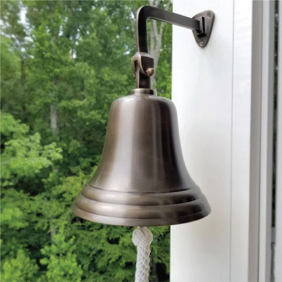 10 inch diameter antiqued brass bell hanging from a wall mount on outside of porch with trees in background