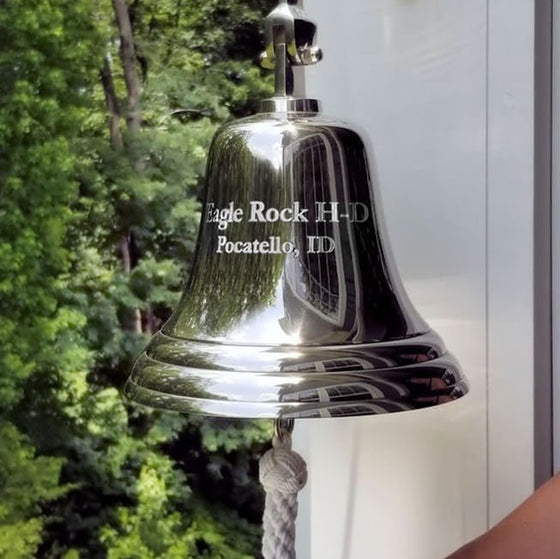 10 inch diameter nickel plated brass wall bell hung outside a porch with two lines of copy engraved  on face of bell and trees in background