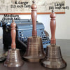 Three different sizes of antiqued brass hand bells with text engraved on them to show differences of size between a nine inch tall, 11 inch and 13 inch versions