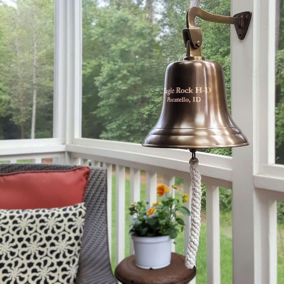 12 inch diameter antiqued finish brass wall bell with two lines of personalized engraving hangs from a wall mount on a back porch