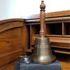 13 inch tall antiqued brass hand bell with contoured wood handle and brass final, plus 3 lines engraving shown on a book with glasses for scale
