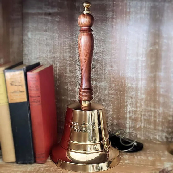 13 inch tall, 6 inch diameter highly polished brass bell with a wood handle on a bookshelf with books and antique reading glasses
