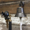 Five inch dark bronze finish brass wall bell with words "Dinner Bell" engraved on front and a candlestick phone nearby for scale