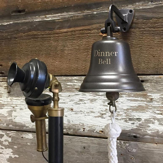 Five inch dark bronze finish brass wall bell with words "Dinner Bell" engraved on front and a candlestick phone nearby for scale