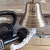 Closeup of 7 inch antiqued brass wall bell with PR BELL engraved on front and black cast iron weights in back