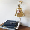 7 inch polished gold finish solid brass bell with red and silver pewter Maltese Cross Firefighter medallion mounted on wall over books and vintage glasses for size reference