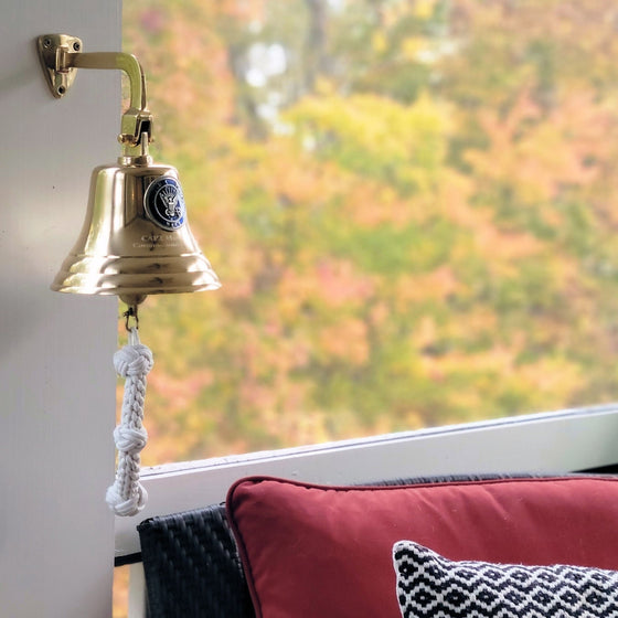 7 inch diameter polished gold solid brass wall bell with blue and silver navy pewter medallion mounted on outdoor porch wall with fall leaves in background