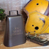 A dark bronze finish brass cowbell with the engraving 'Rx: MORE COWBELL' on its side, placed on a wooden surface with a pair of glasses and a blurred background featuring a yellow antique football helmet